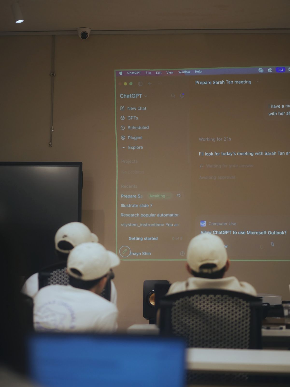 Learners in caps watching a ChatGPT demo projected on the classroom wall